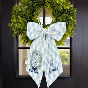 Op de afbeelding: Een decoratieve strik met een blauw-wit geblokt patroon, hangend aan een deur. De strik heeft een monogram met de letter "S" in een krans en de naam "The Smiths" in script. Een groene krans is op de achtergrond te zien.