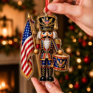 May include: A flat acrylic nutcracker ornament featuring a soldier in a blue, red, and gold uniform with a tall hat. The figure holds a large American flag and a drum. The name Jaycob is printed on the hat brim. It hangs from a red ribbon.