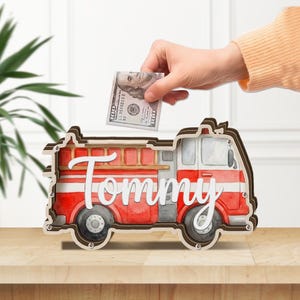 May include: A wooden fire truck-shaped bank with the name "Tommy" in white script. The bank is painted red and white with black wheels and a ladder. A hand is placing a $100 bill into the slot.