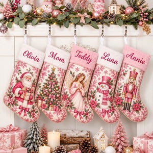 Peut inclure: Cinq chaussettes de Noël avec des revers blancs et des motifs floraux roses. Chaque chaussette présente un personnage unique : le Père Noël, un sapin, un ange, un bonhomme de neige et un casse-noisette. Les prénoms sont brodés sur le revers.