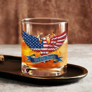 Könnte beinhalten: Ein klares Whiskyglas mit einem patriotischen Design, das einen rot, weiß und blau gefärbten Adler zeigt. Auf dem Glas steht der Text "We The People" auf einem blauen Banner. Das Glas ist mit einer goldenen Flüssigkeit und Eiswürfeln gefüllt.