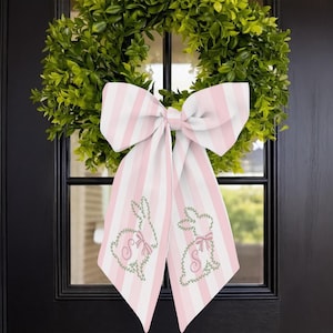 Puede incluir: Una corona de boj verde con un gran lazo a rayas rosas y blancas. El lazo tiene diseños de conejitos bordados con la letra "S" en las colas. La corona está colgada en una puerta negra.