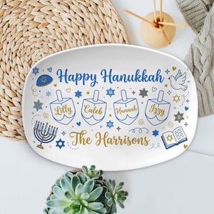 May include: White oval serving platter with blue and gold Hanukkah-themed design. Features the text "Happy Hanukkah" and personalized names on dreidels. Includes menorah, Star of David, and dove illustrations.