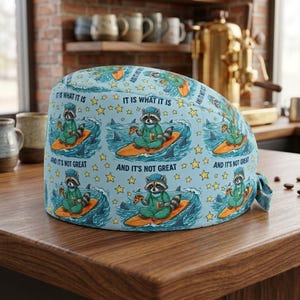 Puede incluir: Un gorro de quir&oacute;fano azul claro con un patr&oacute;n repetitivo de un mapache con uniforme m&eacute;dico, surfeando en una tabla naranja en olas con tiburones, rodeado de estrellas amarillas, con el texto IT IS WHAT IT IS AND IT'S NOT GREAT.