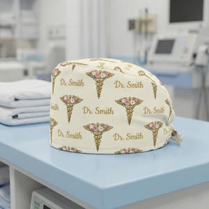 May include: Cream-colored medical scrub cap featuring a repeating pattern of the caduceus symbol with floral accents and the text "Dr. Smith" in gold. The cap is designed for medical professionals.