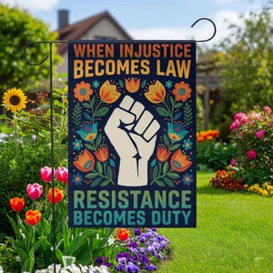 May include: A garden flag with the text "WHEN INJUSTICE BECOMES LAW RESISTANCE BECOMES DUTY" and a white fist graphic. The flag has a dark blue background with floral designs in orange, teal, and white. The flag is displayed in a garden.