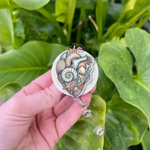 May include: A round badge reel with a heart design incorporating seashells, starfish, and coral. The heart is coloured in blue, tan, and coral, set against a white backdrop. The reel has a retractable cord and a clear plastic clip.