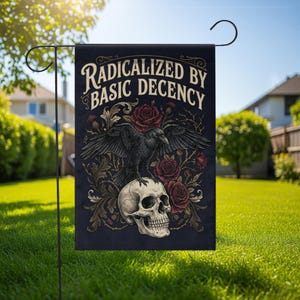 Op de afbeelding: Een zwarte tuin vlag met de tekst "Radicalized by Basic Decency". De vlag toont een raaf die op een schedel zit, omringd door rozen en sierlijke bloemmotieven. De vlag wordt buiten in een zonnige tuin getoond.