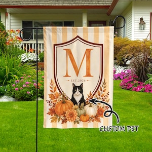 Könnte beinhalten: Eine rechteckige Gartenflagge mit vertikalen beigen Streifen zeigt ein zentrales braunes Wappen mit einem großen orangefarbenen Buchstaben M, dem Text EST.2026 und einer schwarz-weißen Tuxedo-Katze inmitten von orangefarbenen Kürbissen und Herbstlaub.