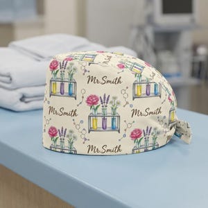 May include: A beige surgical scrub cap with a repeating pattern of flowers, test tubes, and molecular structures. The text "Mr. Smith" is also printed on the cap. The cap is made of a soft, lightweight fabric.