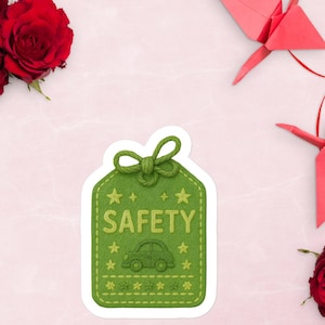 Könnte beinhalten: Ein grüner Aufkleber mit dem Wort "SAFETY" und einer Autografik. Der Aufkleber hat eine Anhängerform mit einer Schleife oben und Stern- und Stichdetails. Rote Rosen und Origami-Kraniche sind im Hintergrund.