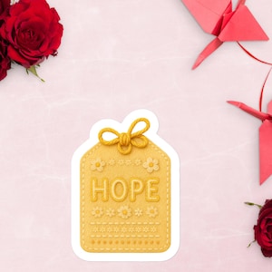 Può includere: Un adesivo giallo con la parola "HOPE" in lettere in rilievo. L'adesivo ha la forma di un'etichetta con un fiocco in alto e dettagli floreali e puntinati. Rose rosse e gru origami sono disposte attorno all'adesivo su uno sfondo rosa.