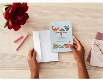 May include: A light blue birthday invitation with dinosaur illustrations and the text "Roaring into 7". The invitation is held near a white envelope, a pink pen, and a red flower.