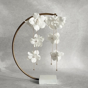May include: A pair of long, dangling earrings featuring multiple white fabric flower petals attached to thin gold-toned chains. Each flower is accented with a small central bead, and the earrings are displayed on a curved, bronze-colored metal stand.