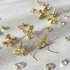 以下が含まれることがあります： 中央に透明なクリスタルのアクセントをあしらった、繊細な金属製の花モチーフのゴールドトーンのドロップピアス。各ピアスはポストバックに取り付けられた細いゴールドチェーンから吊り下げられており、近くには小さな透明なクリスタルビーズが散らばっています。