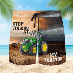 May include: Swim trunks with a green tractor graphic and the text "STOP STARING AT MY TRACTOR". The shorts have a drawstring and a landscape design with a field and stormy sky.