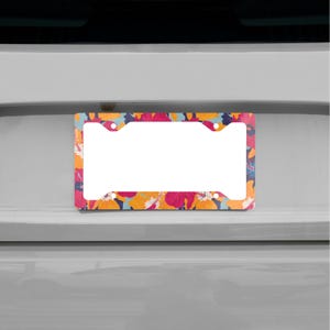Puede incluir: Marco de matrícula con un vibrante estampado floral. El marco presenta un centro blanco para la placa y está adornado con flores coloridas en tonos de rosa, naranja y azul sobre un fondo oscuro. El marco tiene cuatro orificios de montaje.
