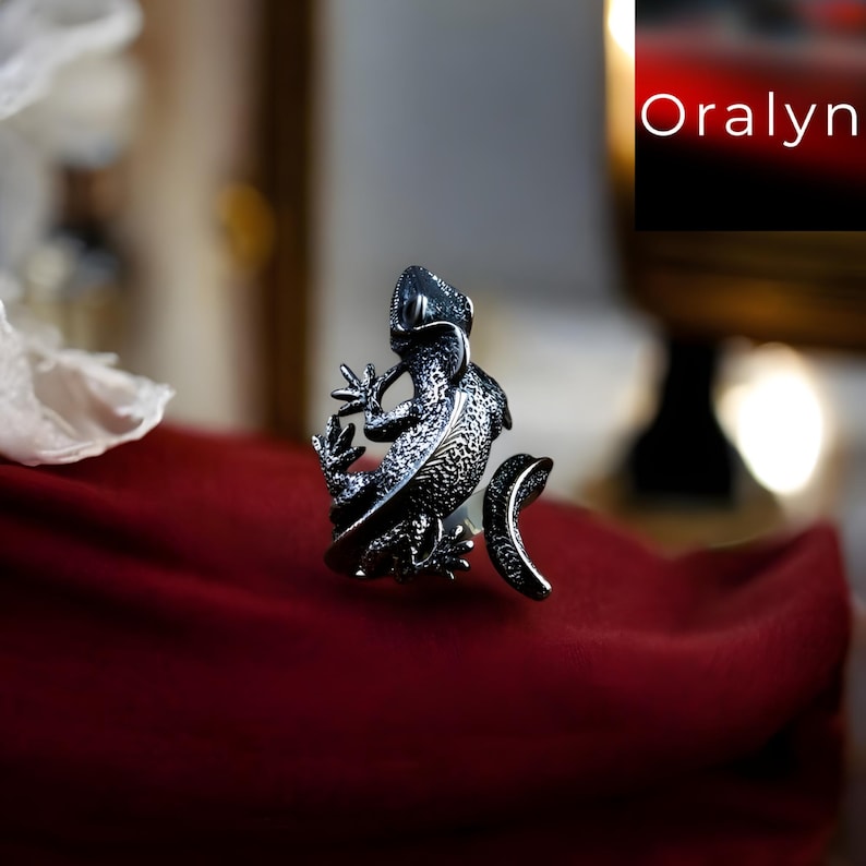 May include: A silver-toned lizard ring with intricate detailing. The ring features a textured lizard design with a curved tail, set against a deep red background. The brand name "Oralyn" is visible in the upper right corner.