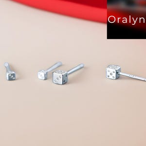 Puede incluir: Pendientes de botón plateados con forma de dados. Cada pendiente presenta un pequeño dado cúbico con marcas de puntos visibles, montado sobre un fino poste. Los pendientes se muestran sobre un fondo neutro, con un acento rojo en la esquina superior.