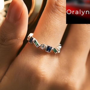 May include: A silver ring with a geometric design featuring blue and green rectangular stones and a small, round, clear stone. The ring is on a finger, with the brand name "Oralyn" visible in the background.