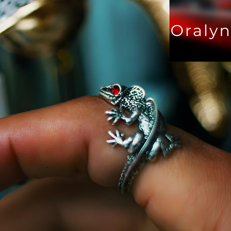 May include: A silver-colored lizard ring with red gemstone eyes. The ring is wrapped around a finger, showcasing the detailed texture of the lizard's scales and limbs. The ring is a unique piece of jewelry.