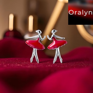 Könnte beinhalten: Ohrstecker mit silbernen Ballerinas. Jeder Ohrring zeigt eine Tänzerin mit rotem Tutu. Die Ohrringe sind vor einem tiefroten Hintergrund platziert. Der Markenname "Oralyn" ist im oberen rechten Eck sichtbar.