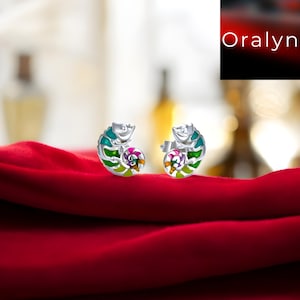 Puede incluir: Un par de pendientes de camaleón plateados con detalles de esmalte colorido. Los pendientes presentan un diseño en espiral con tonos de turquesa, verde, rosa y amarillo. Se exhiben sobre una superficie de satén rojo.