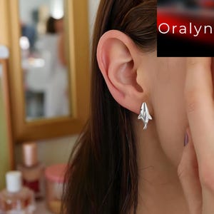 May include: Silver shark stud earrings. The earrings are small, detailed, and feature a shark design. The earrings are shown on an ear, with the focus on the jewelry. The brand name "Oralyn" is visible in the upper right corner.