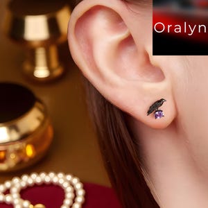 Op de afbeelding: Een zwarte kraai-oorbel met een paarse edelsteen. De oorbel wordt op een oor gedragen. Het woord "Oralyn" staat in de rechterbovenhoek.