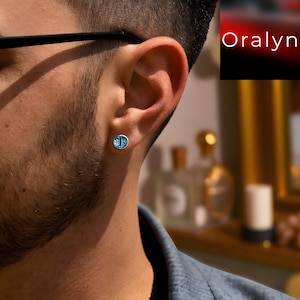 Puede incluir: Un pendiente de botón plateado con un centro redondo, azul claro y estampado se lleva en la oreja. El pendiente se coloca sobre un fondo borroso con la palabra "Oralyn" en la esquina superior derecha.
