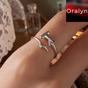Puede incluir: Un anillo abierto de tono plateado con forma de tiburón martillo, con la cabeza del tiburón en un extremo y la cola en el otro. El metal tiene un acabado liso y pulido. El texto "Oralyn" es visible en la esquina superior derecha.