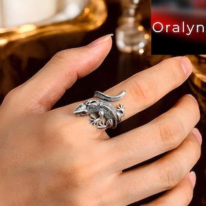 以下が含まれることがあります： 指に巻き付いた、トカゲの形をした銀色のリング。トカゲは詳細な鱗と小さな赤い目をしています。リングは、肌の色の明るい手の上に表示されています。