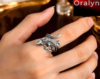 Puede incluir: Un anillo plateado con forma de lagarto, enrollado alrededor de un dedo. El lagarto tiene escamas detalladas y pequeños ojos rojos. El anillo se muestra en una mano de piel clara.