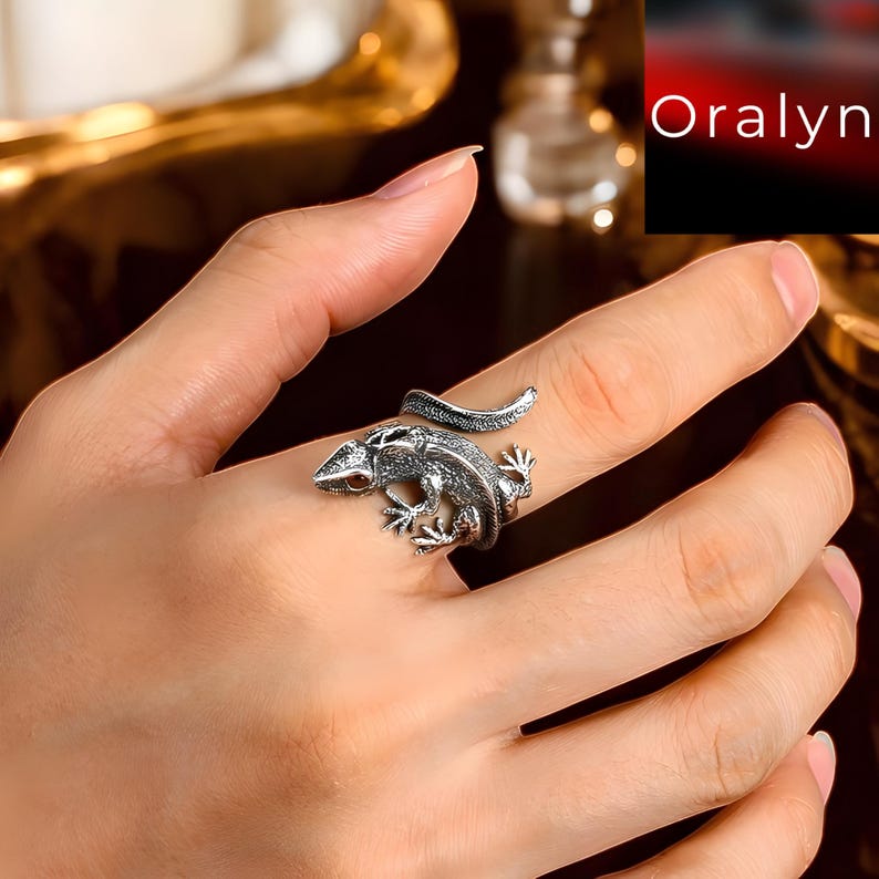 May include: A silver-toned ring in the shape of a lizard, wrapped around a finger. The lizard has detailed scales and small red eyes. The ring is shown on a hand with a light skin tone.