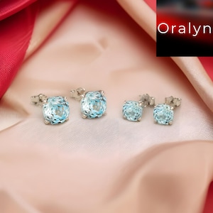 Könnte beinhalten: Vier Paar hellblaue Edelstein-Ohrstecker. Die Ohrringe sind in Silber gefasst und haben einen quadratischen Kissenschliff. Die Ohrringe werden auf einem rosa Satin-Stoff präsentiert.