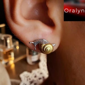 Peut inclure: Une boucle d'oreille en forme d'escargot avec un corps en argent texturé et une coquille dorée lisse et spiralée. La boucle d'oreille est placée sur un lobe d'oreille. Le texte 'Oralyn' est visible dans le coin supérieur droit de l'image.
