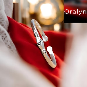 以下が含まれることがあります： オープンデザインのシルバーカラーのカフブレスレット。ブレスレットは装飾的なエッチングパターンと中央のデザイン要素を備えています。背景は鮮やかな赤い生地で、右上隅に「Oralyn」という文字があります。