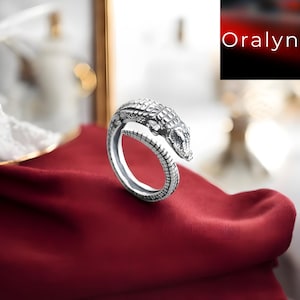 Puede incluir: Un anillo plateado con forma de cocodrilo, con escamas detalladas y un diseño texturizado. La cabeza del cocodrilo se curva para encontrarse con la cola, formando la banda del anillo. El anillo se exhibe sobre una tela roja oscura.