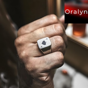 May include: A silver ring with a rectangular face featuring the Ace of Spades playing card design. The ring has the letter "A" on each side of the spade symbol. The ring is shown on a person's hand.