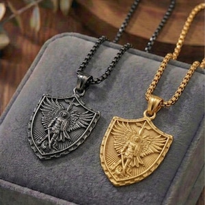 May include: Two shield-shaped pendant necklaces. One is gunmetal-colored, the other gold-toned. Each pendant features a detailed relief of a winged figure with a sword and shield. The necklaces rest on a gray velvet surface.