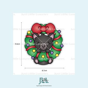 May include: A black cat peeking out of a Christmas wreath. The wreath is green with red ribbon and colorful ornaments. The image is approximately 8.7cm wide and 9.4cm tall. The text "Just Little Luxuries" is visible.