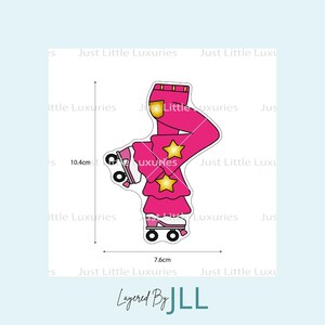 Puede incluir: Un diseño de patines de color rosa con detalles de estrellas doradas, que mide 7,6 cm de ancho y 10,4 cm de alto. El diseño incluye el texto "Layered By JLL" y "Just Little Luxuries".