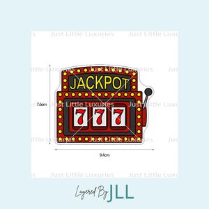 Puede incluir: Un diseño de máquina tragamonedas roja y negra con la palabra "JACKPOT" en la parte superior y los números "777" en las ventanas. El diseño mide 9,4 cm de ancho y 7,6 cm de alto.