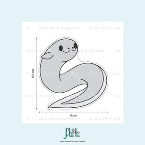 May include: A gray seal-shaped design with a black outline, measuring 9 cm wide and 9.4 cm tall. The design includes small black eyes and a smiling mouth. The text "Just Little Luxuries" is repeated around the design.