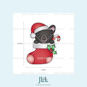 May include: A Christmas-themed koala bear cookie cutter. The koala is wearing a Santa hat and holding a candy cane. It is inside a red stocking with holly. The cutter is 9.4cm tall and 7.6cm wide.