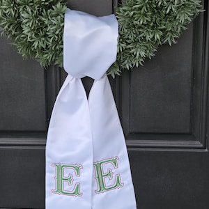 Puede incluir: Faja de puerta de satén blanco con la letra "E" bordada en verde y rosa a cada lado. La faja cuelga de una puerta verde oscuro con una corona verde en el fondo. La faja está atada con un lazo.