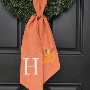 May include: An orange fabric door hanger with a green wreath. The door hanger has a white letter "H" and an embroidered pumpkin. The door hanger is tied to the wreath and hangs on a black door. A seasonal autumn decoration.