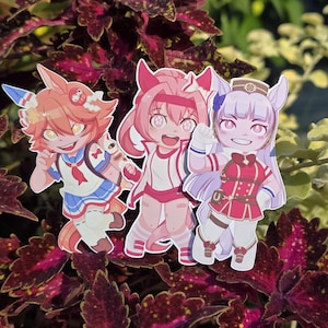 Op de afbeelding: Drie stickers van anime-personages. De personages hebben levendige kleuren, waaronder oranje, roze en paars. Ze dragen gedetailleerde outfits met accessoires. De stickers staan tegen een achtergrond van donkerrood en groen gebladerte.