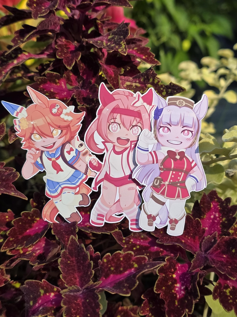 Op de afbeelding: Drie stickers van anime-personages. De personages hebben levendige kleuren, waaronder oranje, roze en paars. Ze dragen gedetailleerde outfits met accessoires. De stickers staan tegen een achtergrond van donkerrood en groen gebladerte.