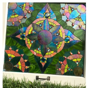 May include: A colorful stained glass window with a geometric design featuring a central flower motif in shades of green, blue, and purple. The window is framed in white wood and set against a backdrop of green grass.